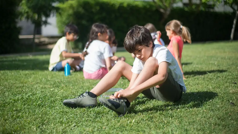 A child tying shoelaces while sitting on grass in a sunny park surrounded by friends.
