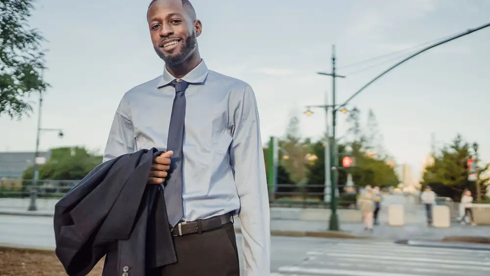 Businessman in formal attire smiling and holding a suit jacket outdoors in an urban setting.