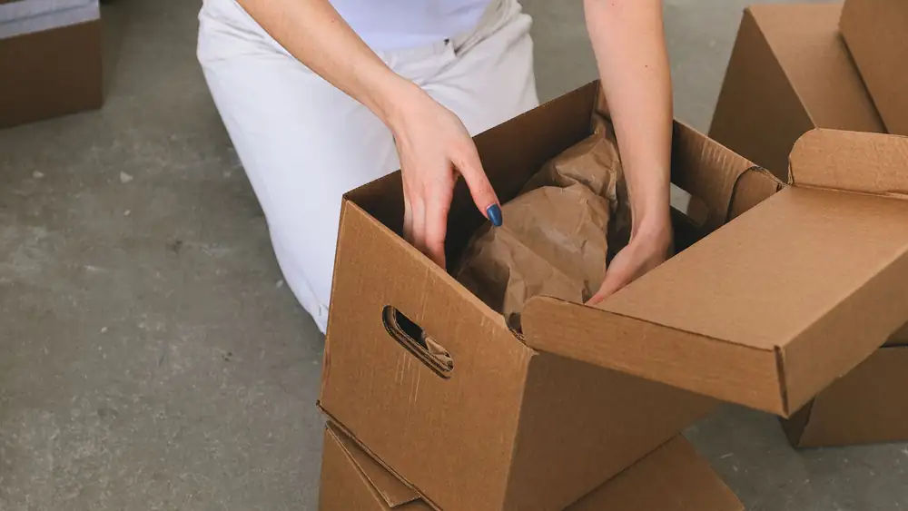 From above of crop anonymous relocating female packing wrapped items into carton box while preparing for moving to new apartment