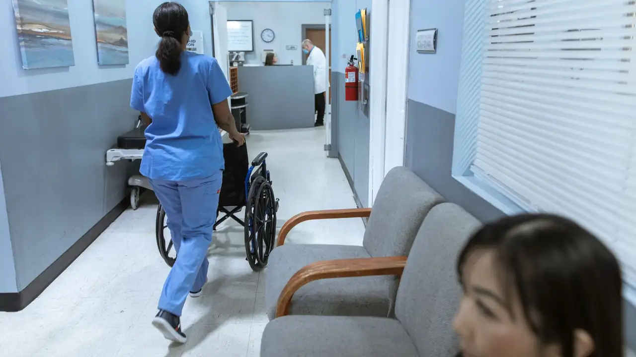 A nurse in scrubs pushes a wheelchair through a well-lit hospital corridor, attending to patient care.