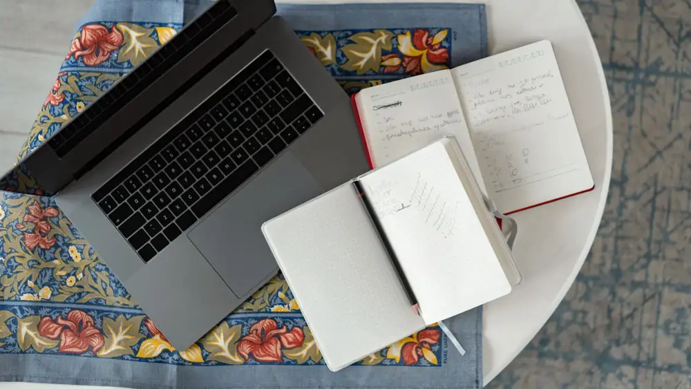 Overhead shot of a laptop with notebooks and a pencil on a patterned cloth.