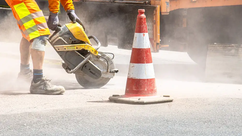 A construction worker cutting asphalt with a large saw next to a traffic cone on a street.